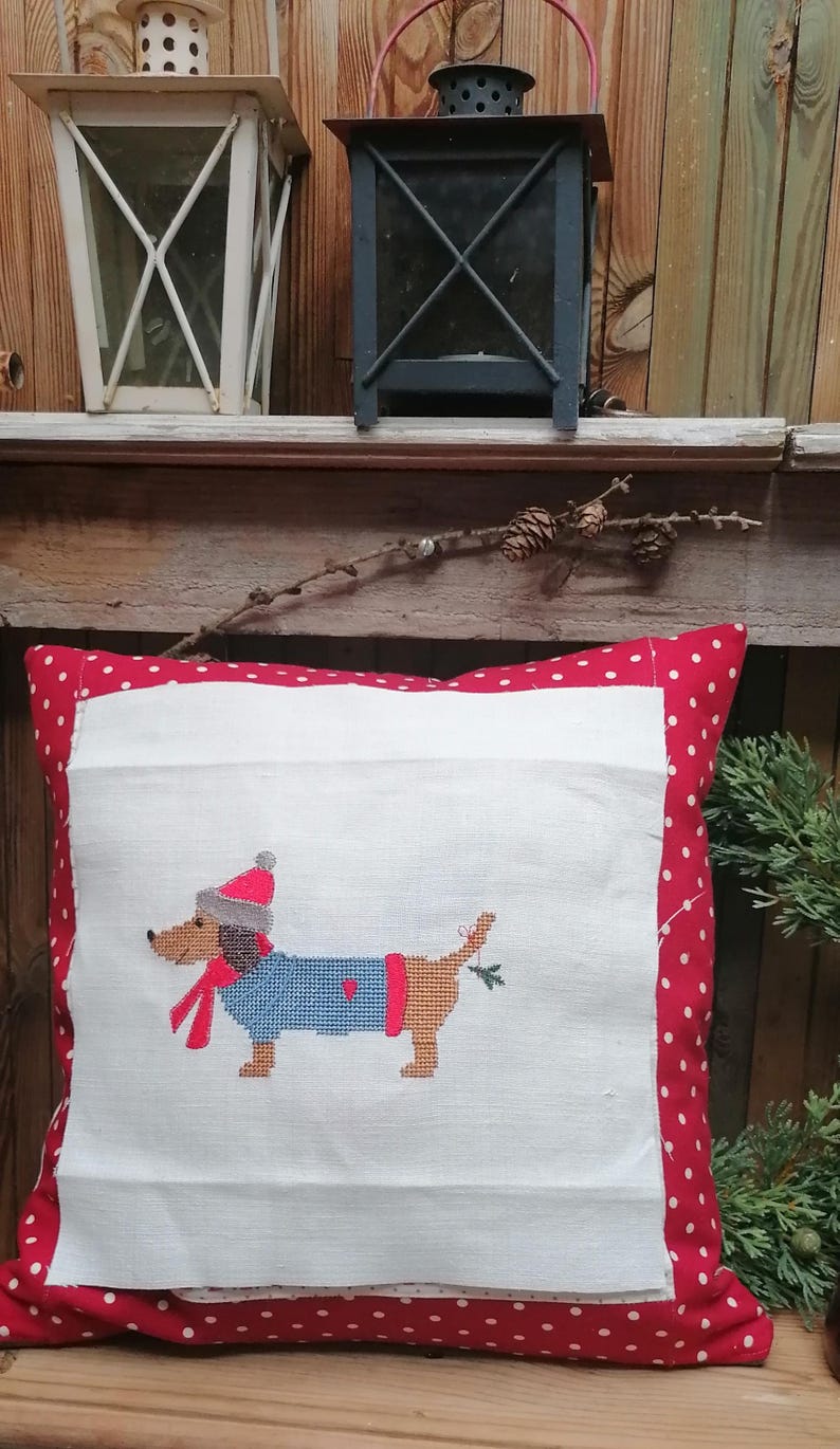 May include: A white pillow with a red polka dot border features a cross-stitch design of a dachshund wearing a blue sweater, red scarf, and a red hat. The dachshund is holding a sprig of holly in its mouth.