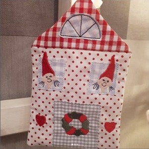 May include: A red and white gingham house-shaped ornament with two embroidered gnomes peeking out of the windows. The house has a wreath on the front and two red hearts on the sides.