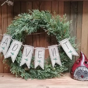 Könnte beinhalten: Ein grüner immergrüner Kranz mit einem Banner aus grobem Leinen, auf dem "Advent" steht. Das Banner ist mit einer Schnur befestigt und auf jedem Fähnchen sind kleine grüne Zweige gestickt. Rechts vom Kranz befindet sich eine kleine rote und graue Gnomfigur.