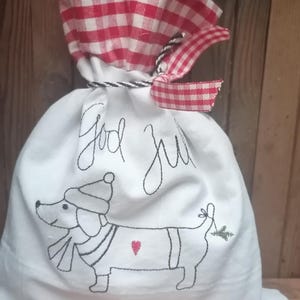 May include: White fabric gift bag with a red and white checkered top. The bag is embroidered with a dachshund wearing a hat and scarf, a heart, and a sprig of greenery. The bag also says "God Jul" in black embroidery.