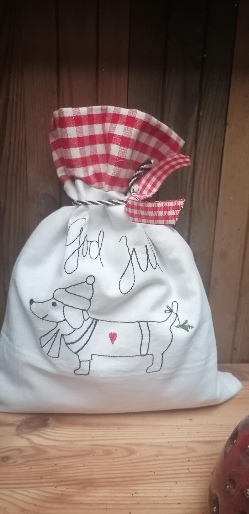 May include: White fabric gift bag with a red and white checkered top. The bag is embroidered with a dachshund wearing a hat and scarf, a heart, and a sprig of greenery. The bag also says "God Jul" in black embroidery.