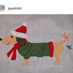 May include: Cross-stitch pattern of a dachshund wearing a green sweater, red scarf, and a Santa hat. The dog is holding a sprig of holly in its mouth.