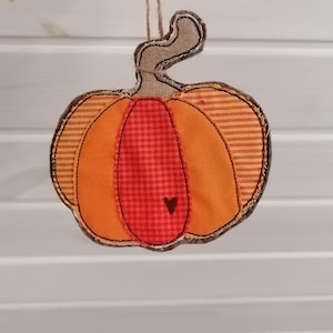 Könnte beinhalten: Handgefertigte Stoff-Kürbis-Dekoration mit orangefarbenen und roten Akzenten. Der Kürbis hat eine braune Umrandung und ein kleines Herzdetail. Aufgehängt an einer natürlichen Schnur, perfekt für die Herbstdekoration.