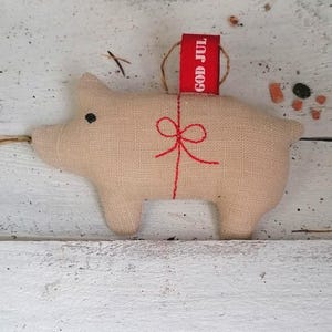 Op de afbeelding: Een beige linnen varkensornament met een rood geborduurd strikje en een rood label met de tekst "God Jul".
