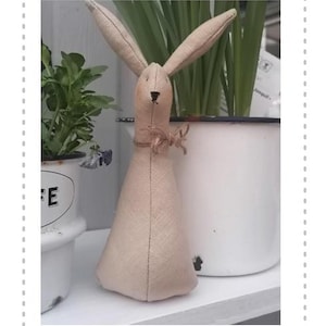 May include: A beige fabric bunny rabbit with long ears and a brown fabric bow around its neck. The bunny is sitting on a white surface.