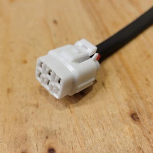 May include: A white electrical connector with multiple pin receptacles, attached to a black wire. The connector has a rectangular shape with a locking mechanism. The background is a wooden surface.