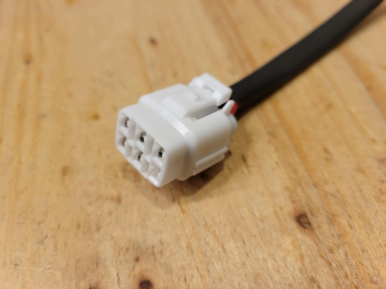 May include: A white electrical connector with multiple pin receptacles, attached to a black wire. The connector has a rectangular shape with a locking mechanism. The background is a wooden surface.