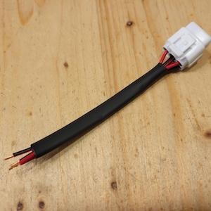 May include: Electrical wiring harness with a white connector and black insulation. The harness has red and black wires extending from the connector and exposed wire ends. The background is a light brown wooden surface.