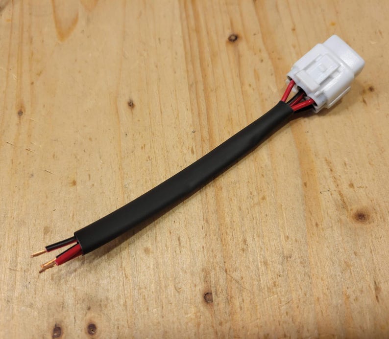 May include: Electrical wiring harness with a white connector and black insulation. The harness has red and black wires extending from the connector and exposed wire ends. The background is a light brown wooden surface.