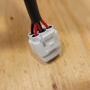 May include: Close-up of an electrical connector with a white plastic housing. The connector has multiple red and black wires emerging from it, encased in a black insulated cable. The background is a light brown wooden surface.