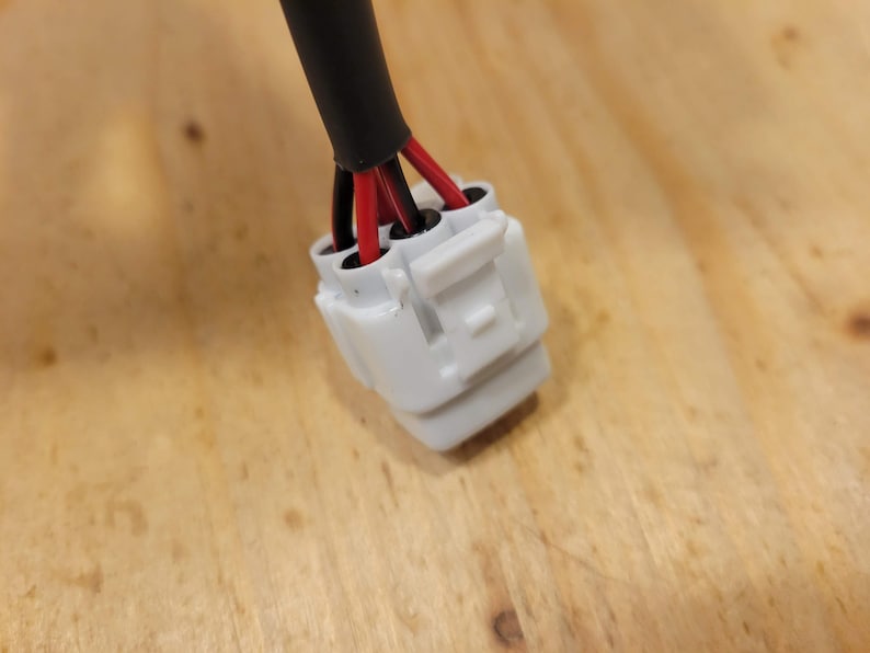 May include: Close-up of an electrical connector with a white plastic housing. The connector has multiple red and black wires emerging from it, encased in a black insulated cable. The background is a light brown wooden surface.