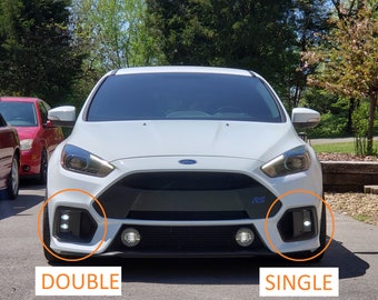 Puede incluir: Un Ford Focus RS blanco con un parachoques delantero negro y una sola luz antiniebla redonda en el lado derecho y dos luces antiniebla redondas en el lado izquierdo. El coche está aparcado en una entrada pavimentada.