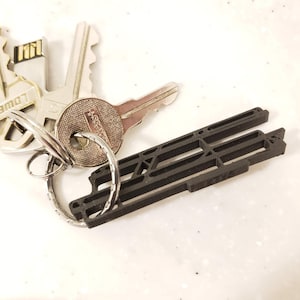 May include: A set of keys on a silver key ring, including a USB key and a black, geometric keychain. The keys are silver and have various shapes and markings. The keychain is a dark color and has a unique, architectural design.