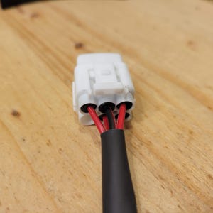 May include: A white electrical connector with three black holes, each containing a red and black wire. The wires are encased in a black insulated sleeve. The connector is on a wooden surface.