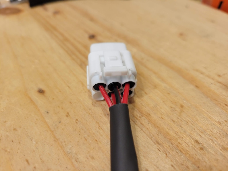 May include: A white electrical connector with three black holes, each containing a red and black wire. The wires are encased in a black insulated sleeve. The connector is on a wooden surface.