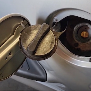 May include: A close-up of a silver gas cap with a black plastic lever. The gas cap is open and the lever is in the up position.