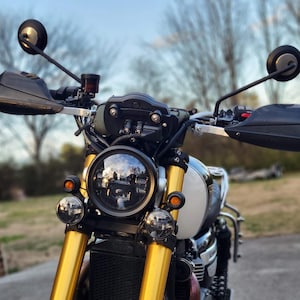 Puede incluir: Primer plano de una motocicleta con manillares dorados y protectores de manos negros. La motocicleta tiene un faro redondo y dos luces redondas más pequeñas a cada lado. La motocicleta está estacionada en una carretera pavimentada.