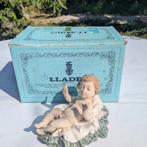 Può includere: Statuina in porcellana di un cherubino sdraiato su un letto di tessuto verde e bianco. Il cherubino ha la pelle chiara e i capelli biondi. Una scatola blu con la scritta "LLADRO" è dietro la statuina.