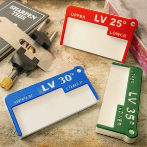 May include: A collection of sharpening guides in red, blue, and green, each with a white interior. The guides are labeled with angles: LV 25°, LV 30°, and LV 35°. The text "UPPER" and "LOWER" are also visible. A sharpening tool and a book with the text "SHARPEN THIS" are also in the image.