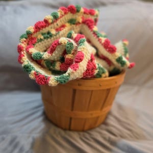 May include: A crocheted blanket with a cream-coloured base featuring a decorative border of alternating pink and green bobble stitches. The blanket is folded and placed inside a round, light brown wooden basket with vertical slats and metal staples.