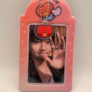 May include: Pink photo ID holder with a photo of a person waving. The holder has a cartoon heart-shaped character charm attached. The holder has a paw print pattern and a rounded top.