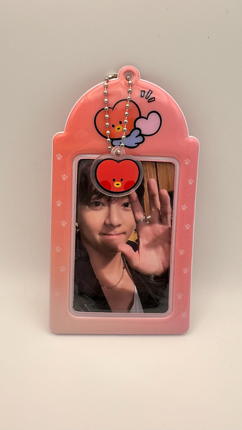 May include: Pink photo ID holder with a photo of a person waving. The holder has a cartoon heart-shaped character charm attached. The holder has a paw print pattern and a rounded top.