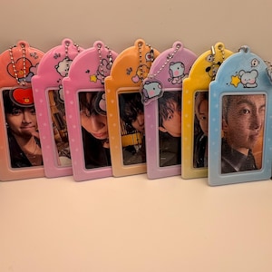 Op de afbeelding: Een verzameling kleurrijke fotokaarthouders in verschillende pasteltinten, waaronder roze, oranje, geel en blauw. Elke houder heeft een stripfiguur en een ketting. De houders bevatten foto's van mensen.