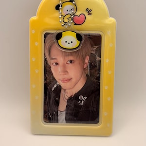 May include: A yellow photo card holder with a cartoon character charm at the top. The holder contains a photo of a person. The holder has a paw print design on the sides.