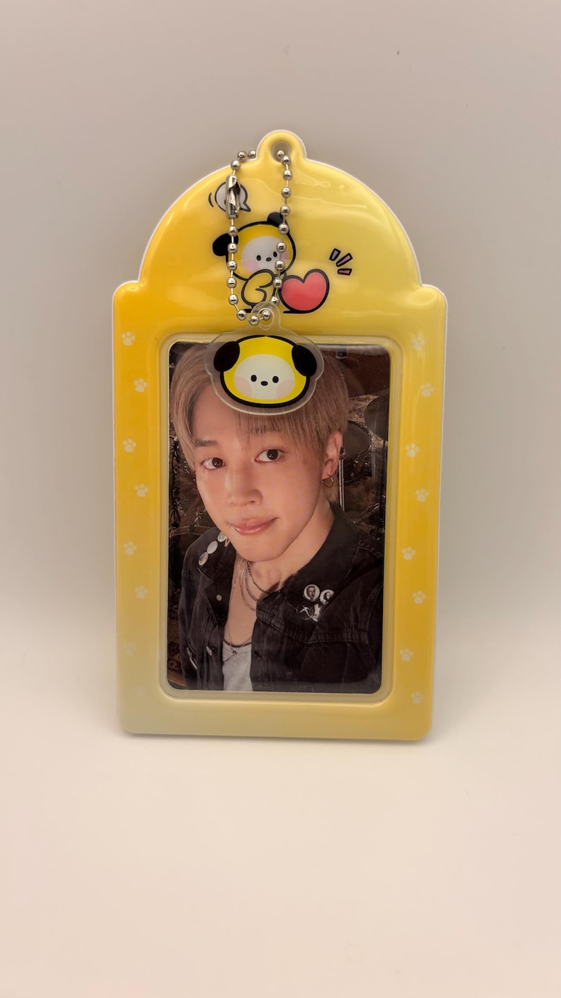 May include: A yellow photo card holder with a cartoon character charm at the top. The holder contains a photo of a person. The holder has a paw print design on the sides.