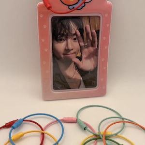 May include: A pink photo holder with a cartoon character and a red cord, holding a photo of a person waving. Below are colorful keychains in red, yellow, blue, pink, and green.