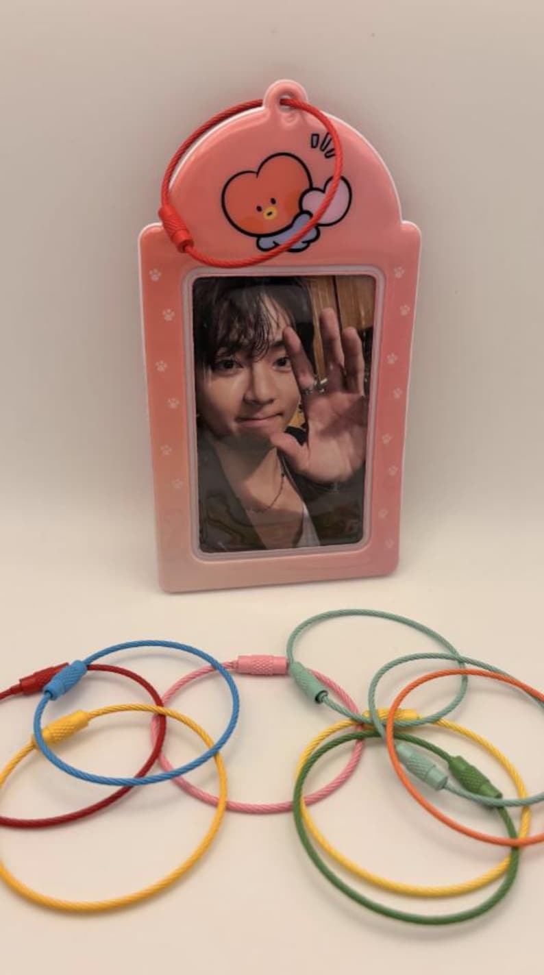 May include: A pink photo holder with a cartoon character and a red cord, holding a photo of a person waving. Below are colorful keychains in red, yellow, blue, pink, and green.