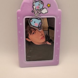 May include: A light purple photo holder with a photo of a person. The holder has a cartoon character charm and paw print details. A silver chain connects the charm to the holder.