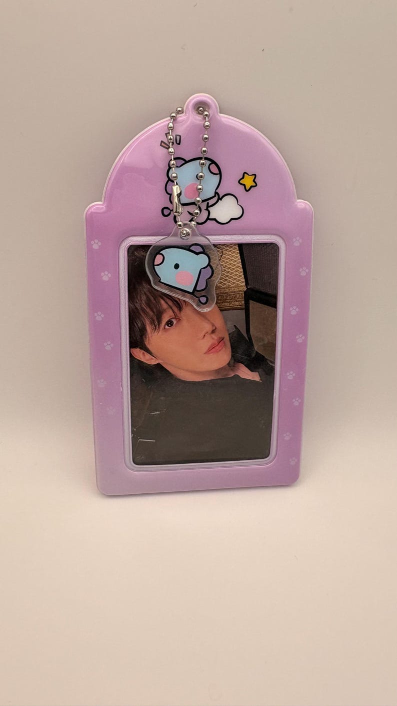 May include: A light purple photo holder with a photo of a person. The holder has a cartoon character charm and paw print details. A silver chain connects the charm to the holder.