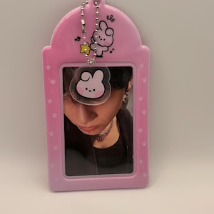 May include: A pink photo card holder with a cartoon bunny design and a silver chain. The holder contains a photo of a person. The holder has a paw print pattern and musical notes.