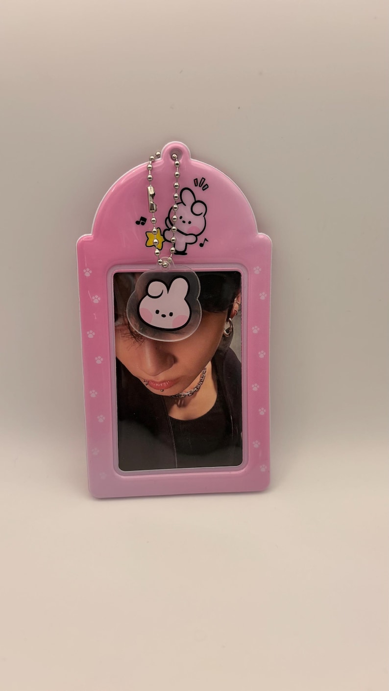 May include: A pink photo card holder with a cartoon bunny design and a silver chain. The holder contains a photo of a person. The holder has a paw print pattern and musical notes.