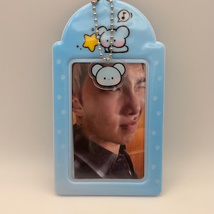 May include: A light blue photo card holder with a photo of a person. The holder has a cartoon bear design and a silver chain. The holder is decorated with paw prints.