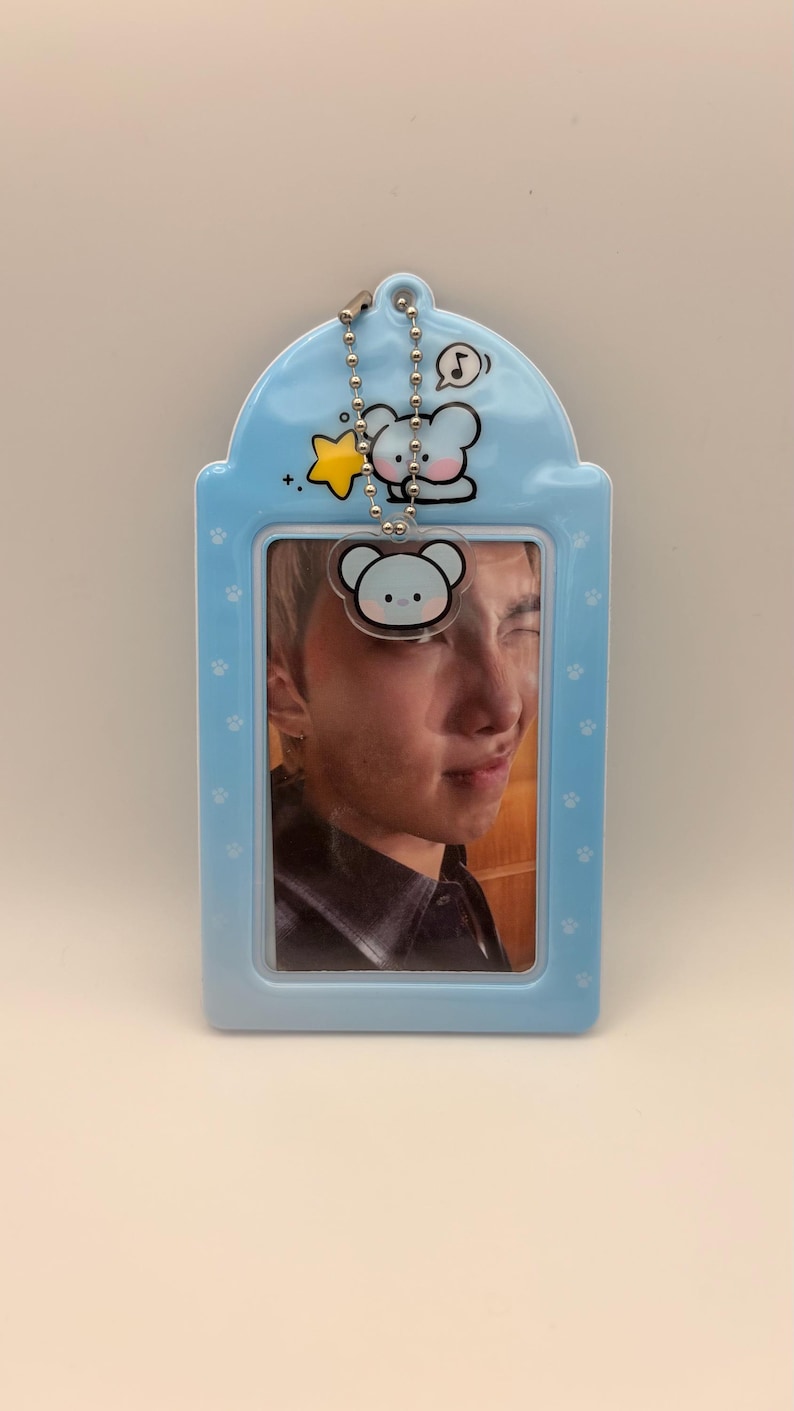 May include: A light blue photo card holder with a photo of a person. The holder has a cartoon bear design and a silver chain. The holder is decorated with paw prints.