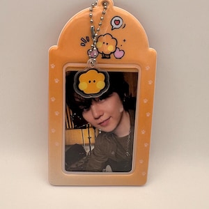May include: Orange keychain with a photo of a person. The keychain has a rectangular shape with rounded edges and paw prints. A small charm with a cartoon face and a heart is attached to the top.