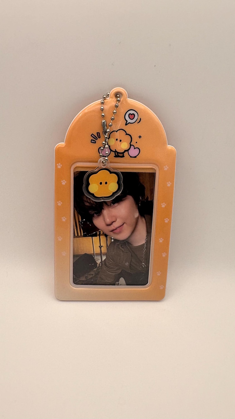 May include: Orange keychain with a photo of a person. The keychain has a rectangular shape with rounded edges and paw prints. A small charm with a cartoon face and a heart is attached to the top.