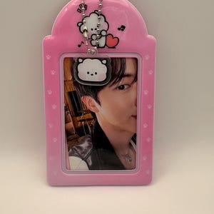 May include: Pink ID badge holder with a cartoon bear design and a photo of a person. The holder has a chain and paw print details. The photo shows a person wearing a black jacket and a necklace.