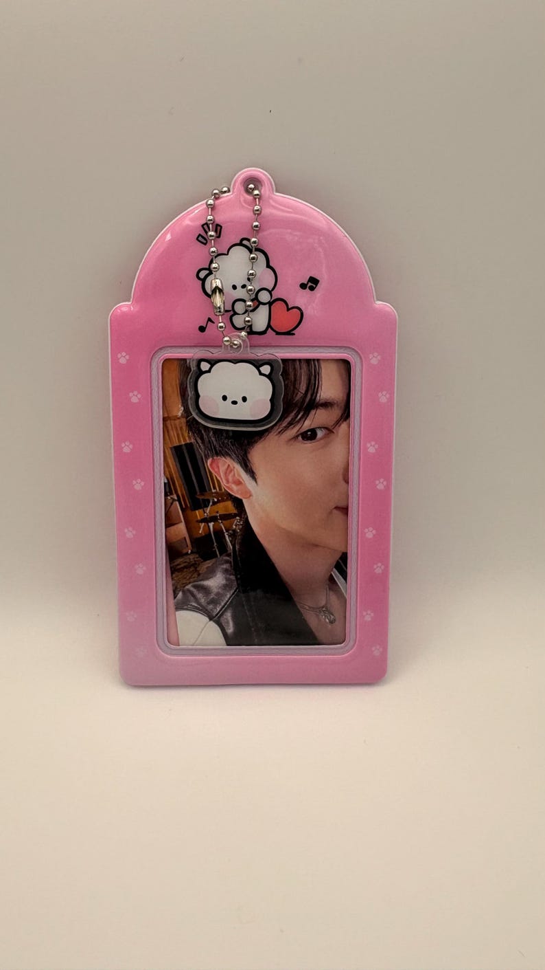 May include: Pink ID badge holder with a cartoon bear design and a photo of a person. The holder has a chain and paw print details. The photo shows a person wearing a black jacket and a necklace.