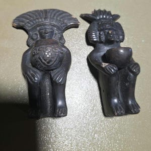 May include: Two dark gray, small statues of figures with elaborate headdresses. One figure holds a vessel, the other a bowl. The statues have detailed facial features and are in a seated position. The statues appear to be made of a stone-like material.