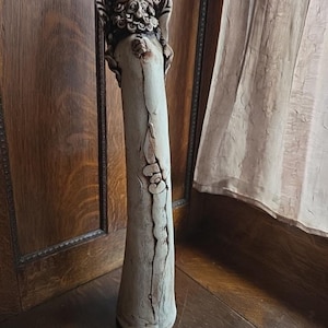 May include: A tall, off-white ceramic vase with a detailed face and headdress at the top. The vase has a cylindrical body with a textured surface and a crack running down the front. The base is wider than the top.