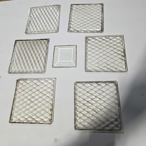 Può includere: Una collezione di piastrelle di vetro trasparente con una superficie strutturata a motivo di diamante. Le piastrelle sono quadrate e rettangolari, con una piastrella quadrata più piccola al centro. Il vetro ha una qualità riflettente e i bordi sono incorniciati.