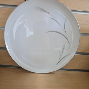 Op de afbeelding: Een witte keramische plaat met een zilveren rand en een tarwe-aarontwerp in grijs. De plaat is rond en heeft een glanzende afwerking. Het tarwe-aarontwerp bevindt zich aan de linkerkant.