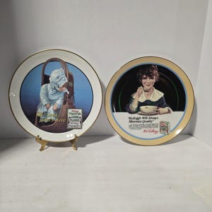 Op de afbeelding: Twee vintage decoratieve borden. Eén bord toont een baby in een mand met de tekst "Oh! Look who's here" en "Kellogg's Toasted Corn Flakes". Het andere bord toont een kind dat ontbijtgranen eet met de tekst "Kellogg's Will Always Maintain Quality".