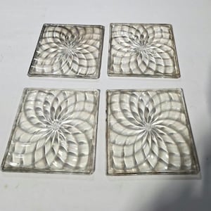 May include: Four clear glass square coasters with a swirling floral design. The coasters are made of a thick glass material and are designed to protect surfaces from water rings and heat.