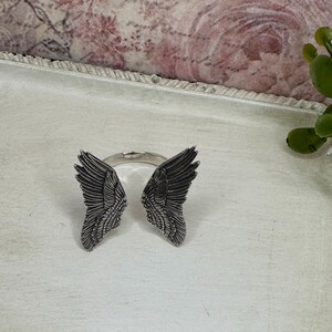 Op de afbeelding: Een zilveren ring met aan weerszijden twee engelenvleugels. De vleugels zijn gedetailleerd en hebben een textuur.