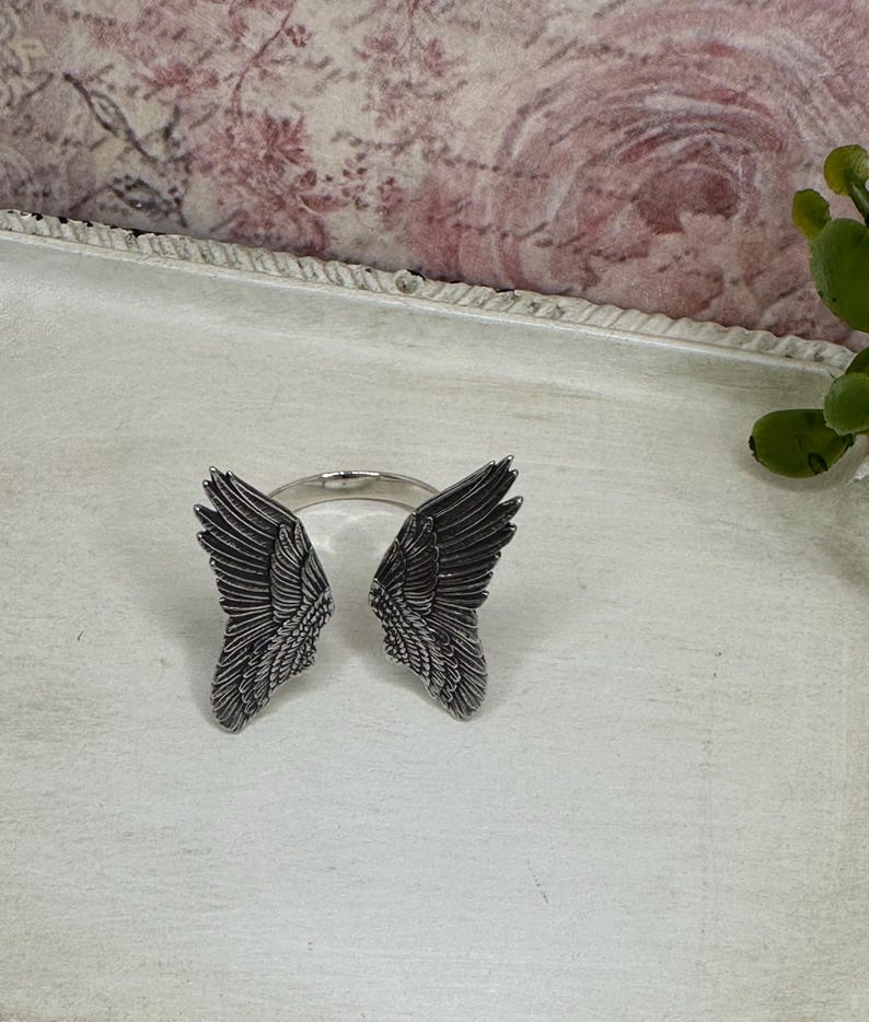 Op de afbeelding: Een zilveren ring met aan weerszijden twee engelenvleugels. De vleugels zijn gedetailleerd en hebben een textuur.