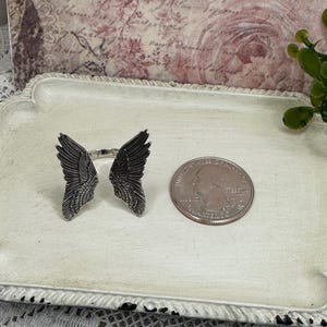 Op de afbeelding: Een zilveren ring met twee engelenvleugels aan weerszijden. De ring ligt op een wit dienblad met een Amerikaanse kwart voor de schaal.